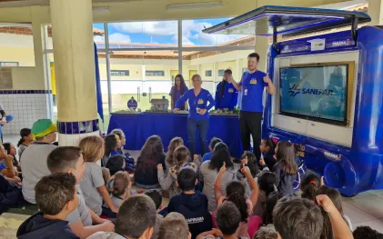 Monitores da Sanepar gesticulam alegremente durante uma palestra educativa sobre saneamento para crianças sentadas no pátio da Escola Maria Nagib, em Guapirama. Ao lado, há uma van adaptada com uma grande tela de TV.