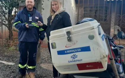 O homem veste o uniforme completo da Sanepar/Enorsul e segura uma máquina de leitura portátil (leitor de hidrômetro). A mulher loira está ao lado dele, segurando o smartphone