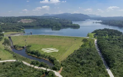 Vista aérea da Barragem de Piraquara com o reservatório de água ao fundo. No gramado do talude da barragem, destaca-se o logotipo estilizado da Sanepar em cor branca