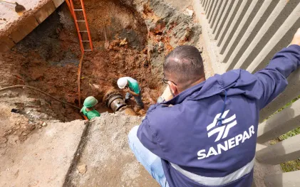 Equipe técnica trabalha na manutenção de uma grande tubulação de água dentro de uma escavação na terra. Em primeiro plano, um supervisor de costas, vestindo jaqueta azul escura com a marca Sanepar, observa o serviço