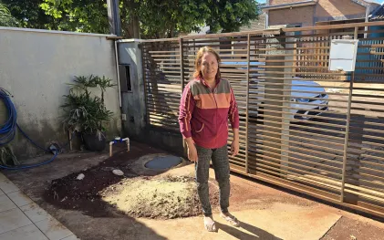 Mulher de casaco colorido e calça escura posa em pé no quintal de sua residência no Jardim Planalto. Ela está ao lado de uma tampa de inspeção de esgoto no chão, com terra onde a fossa foi aterrada. Ela está em frente a um portão de barras horizontais