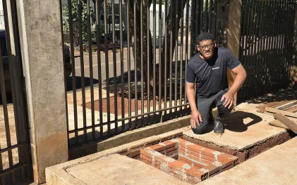Morador sorri ajoelhado na calçada ao lado de uma caixa de inspeção ou passagem de esgoto recém-construída em alvenaria de tijolos. Ele está orgulhoso, pois foi ele quem fez a ligação de esgoto da sua casa. Ao fundo, uma grade metálica preta delimita o espaço residencial