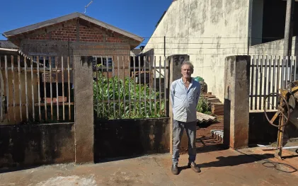 Homem de camisa clara e calça jeans posa em pé na calçada em frente ao portão de uma residência no Jardim Delgado. Ao fundo, aparecem casas de alvenaria sob um céu azul limpo