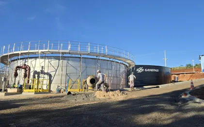 Dois operários trabalham no canteiro de obras de uma estação de tratamento de esgoto. Um deles opera uma betoneira móvel perto de um grande tanque metálico prateado com passarela superior. Ao fundo, um tanque preto exibe o logotipo da Sanepar sob o céu azul