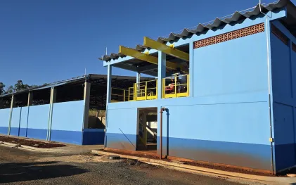 Prédio operacional com pé direito bem alto com paredes pintadas em tons de azul claro e escuro em uma estação de tratamento de esgoto. Anexo uma estrutura possui uma área aberta no piso superior com guarda-corpos amarelos e vigas metálicas, onde estão equipamentos do tipo centrífugas