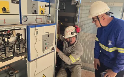 Dois técnicos trabalham em um painel elétrico. Um deles, agachado, realiza ajustes nos cabos internos utilizando capacete com lanterna e protetor auricular, sob a supervisão do colega de uniforme azul