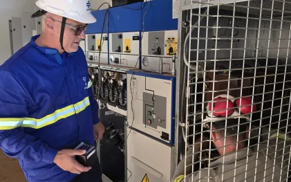 Técnico da Sanepar com uniforme azul e capacete branco observa outro profissional que realiza manutenção interna em um painel elétrico de alta tensão protegido por grade metálica