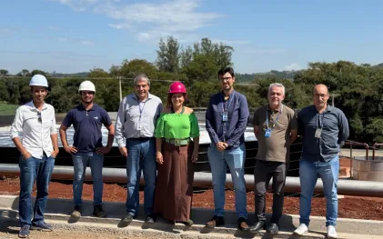 Prefeita e membros da prefeitura, com equipe técnica da Sanepar posam para foto alinhados lado a lado no canteiro de obras da estação de tratamento de esgoto. A prefeita, ao centro, veste blusa verde e capacete rosa, enquanto os demais profissionais usam crachás, camisas casuais e capacetes brancos de proteção