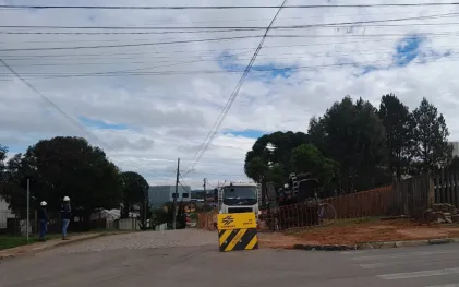 Foto de uma rua em obras com um caminhão branco ao fundo e uma placa amarela da Sanepar em destaque no centro, indicando interdição. Duas pessoas com capacetes brancos observam o local à esquerda.