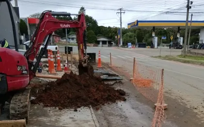 Mini escavadeira vermelha da marca Kubota realizando obras em uma calçada de rua. Há uma pilha de terra escura, cones de sinalização laranjas e uma rede de proteção laranja isolando a área. Ao fundo, vê-se um posto de combustíveis sob um céu nublado.