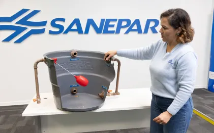 Mulher demonstra os componentes internos de uma caixa d'água utilizando uma maquete em corte que expõe a boia e as tubulações de entrada e saída. Ao fundo, parede branca com o logotipo azul da Sanepar.
