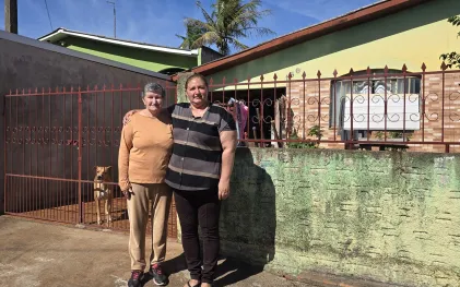 mãe e filha abraçadas na frente de casa, uma senhor com cabelos claros e curtos e a filha sorrindo. no fundo a cachorra caramelo aparece no portão