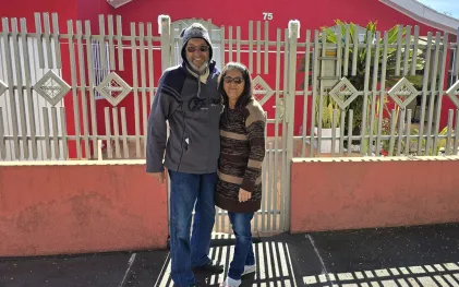 casal em frente ao seu imóvel, ele alto e vestido roupas de lã, incluindo um gorro, ela bem mais baixa abraçada