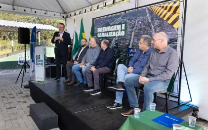 Homem discursa em púlpito com logotipo de Quatro Barras ao lado de painel sobre drenagem e canalização. Autoridades assistem sentadas no palco, onde há uma mesa com uma pasta azul da Sanepar.