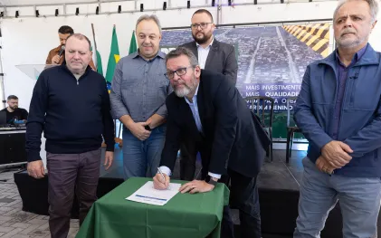 Homem de óculos e terno escuro inclina-se para assinar um documento sobre uma mesa com toalha verde. Ao seu redor, outros quatro homens acompanham o ato em um palco sob uma tenda. Ao fundo, há bandeiras e um banner com a imagem aérea de um bairro.