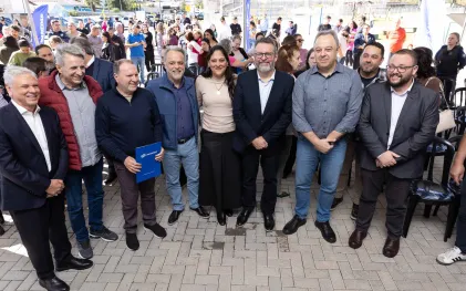 Grupo de autoridades e representantes da Sanepar posa sorrindo para foto em um evento público ao ar livre. Um dos homens segura uma pasta azul com o logotipo da Sanepar, enquanto uma grande quantidade de pessoas acompanha o evento ao fundo.