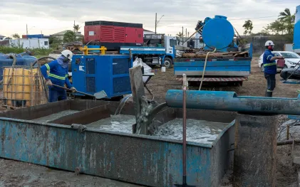Tubulação despejando fluido cinzento de perfuração em um tanque de contenção de metal, com trabalhadores da equipe técnica operando ao redor.