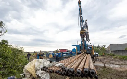 Canteiro de obras para perfuração de poço, com tubos metálicos de perfuração empilhados em primeiro plano e uma grande torre perfuratriz azul ao fundo sob céu nublado.