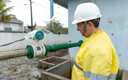 Técnico com capacete branco e capa de chuva amarela observa atentamente o visor de um medidor de vazão digital acoplado a uma tubulação verde.