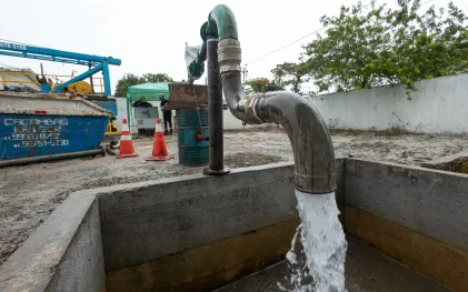 Água jorrando com abundância de uma tubulação metálica para dentro de um tanque de concreto. Ao fundo, dois trabalhadores com capacetes operam equipamentos de perfuração em um canteiro de obras.