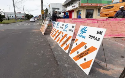 Cavaletes de sinalização com listras brancas e laranjas, logotipo da Sanepar e o texto "EM OBRAS", bloqueiam parte da via, isolando a área onde trabalhadores e maquinário atuam ao fundo.