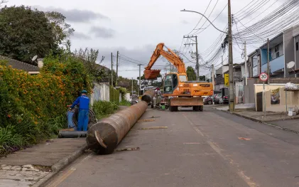 Escavadeira laranja ergue um longo tubo metálico no meio de uma rua residencial, com operários de uniforme azul acompanhando a movimentação da estrutura.