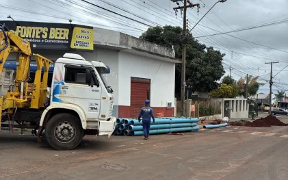 numa rua, um caminhão aparece no canto da foto, no chão ao lado, tem uma pilha de tubulações de água em PVC na cor azul e tem também um trabalhador da obra vestido de azul