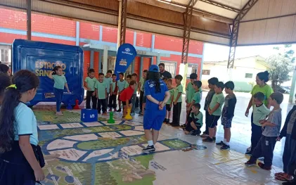 Alunos em fila participando de um jogo de tabuleiro gigante e interativo no chão, mediado por monitores da Sanepar em uma quadra coberta escolar.