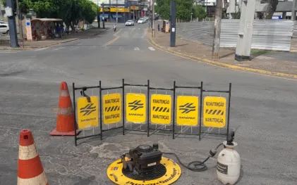 Equipamento de aplicação de fumaça posicionado sobre o poço de visita na rua, isolado por gradis amarelados de sinalização da Sanepar com a palavra "Manutenção" e cones de trânsito