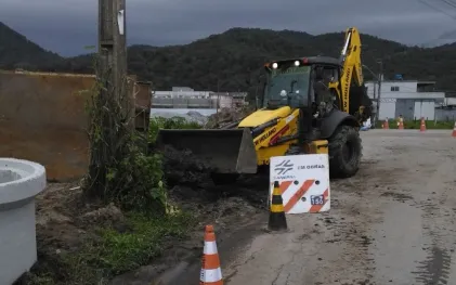 Retroescavadeira amarela movimenta terra na lateral de uma rua asfaltada, próxima a peças circulares de concreto. Cones de trânsito laranjas e uma placa da Sanepar com o aviso "Em obras" e o telefone 156 sinalizam o local.