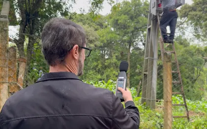 Fotografia em plano médio focada nas costas de um técnico da Sanepar, um homem de cabelos grisalhos e óculos, que segura um decibelímetro digital apontado para cima para medir a intensidade do som. Ao fundo, após uma cerca de arame, outro operador trabalha subido em uma escada de madeira apoiada contra uma estrutura de concreto alta, realizando ajustes em uma caixa de comandos eletrônicos. O ambiente ao redor é repleto de mata verde densa.