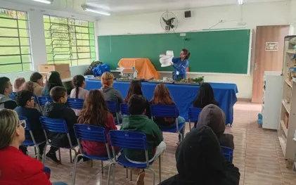 Educadora da Sanepar em sala de aula segura maquete de vaso sanitário durante palestra de conscientização sobre saneamento para alunos