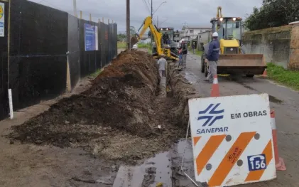 Vala extensa escavada na margem de uma rua asfaltada para a passagem de tubulação. Mais adiante, operários e retroescavadeiras dão andamento à obra. Em primeiro plano, há uma placa de sinalização da Sanepar com os dizeres "Em obras" e o telefone 156.