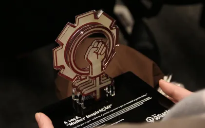 Troféu do Prêmio Mulher InovAÇÃO, em formato de engrenagem com um punho fechado ao centro. Ele é dourado e tem uma base quadrada preta.