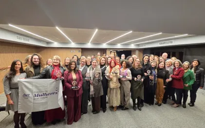 Grupo de mulheres alinhadas em três fileiras dentro de uma sala na sede do Senge-PR. Elas seguram uma bandeira branca do coletivo de mulheres do sindicato.