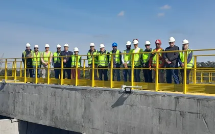 Uma equipe de quinze profissionais e autoridades, vestindo capacetes de segurança e coletes refletivos, posa enfileirada sobre a passarela com guarda-corpo amarelo de uma estrutura de concreto na Estação de Tratamento de Esgoto