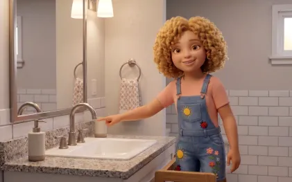Uma menina loira de cabelos crespos e volumosos, vestindo uma jardineira jeans decorada com desenhos de sol e flores sobre uma camiseta rosa, sorri para a câmera em um banheiro. Ela está em pé ao lado de uma bancada de mármore e aponta com o dedo indicador para uma torneira metálica de bica alta que está fechada, incentivando a economia de água. O reflexo de suas costas e do banheiro é visível no espelho da parede.