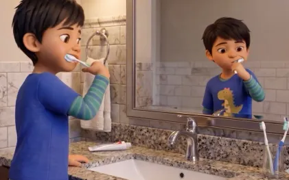 Um menino de cabelo curto e camiseta azul escuro com estampa de dinossauro está escovando os dentes em um banheiro iluminado. Ele está posicionado de perfil à esquerda, olhando para o seu próprio reflexo em um grande espelho à sua frente. A pia de mármore cinza está limpa e a torneira cromada permanece totalmente fechada enquanto ele escova os dentes.