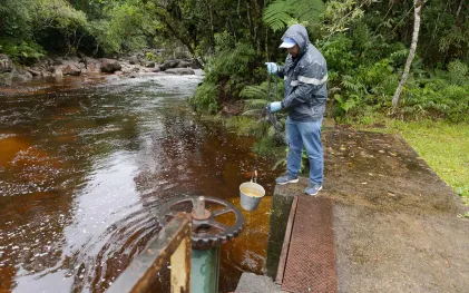 Técnico com capa de chuva e luvas azuis utiliza um balde preso a uma corda para coletar amostra de água em manancial cercado por vegetação.