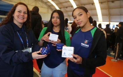 Uma empregada da Sanepar e duas estudantes seguram pequenos coletores de óleo usado em formato de caixa d'´água, redonda e azul, distribuídos pela Sanepar.