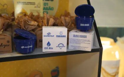 Dois coletores de óleo de cozinha usado em formato de caixa d'água, redondos e da cor azul escura, são exibidos ao lado de suas embalagens, de papelão e brancas. Os itens estão alinhados e possuem a logo da Sanepar