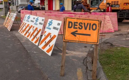 Placa de sinalização amarela com uma seta para a esquerda e o texto "DESVIO". À esquerda, cavaletes zebrados em laranja e branco com a inscrição "OBRAS SANEPAR" delimitam a via interditada. Ao fundo, operários com capacetes e uma escavadeira atuam na obra.