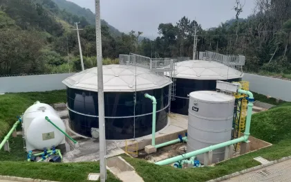 Vista aérea de dois grandes tanques pretos cilíndricos com coberturas metálicas circulares, ladeados por tanques menores e tubulações verdes claros em uma área gramada.