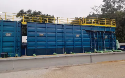 Uma grande estrutura modular metálica pintada de azul-escuro com guarda-corpos amarelos no topo, exibindo tubulações horizontais verdes instaladas na base de concreto.
