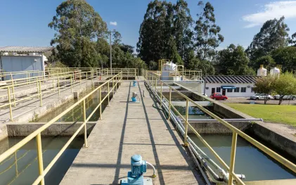 Passarela central da estação de tratamento com motores azuis de bombeamento, ladeada por tanques de água e estruturas operacionais, com árvores e prédios administrativos ao fundo.