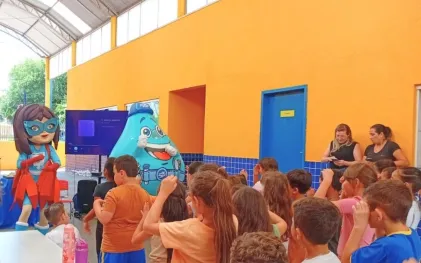 Crianças reunidas em frente a um telão assistindo a uma apresentação com os mascotes da Sanepar: um boneco em formato de gota d'água azul sorridente e uma personagem fantasiada de super-heroína.