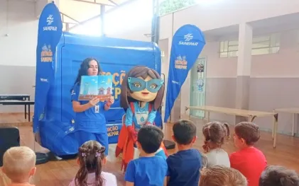 Crianças sentadas no chão assistindo atentamente a uma contação de histórias feita por uma funcionária da Sanepar e pela mascote super-heroína, em frente a uma estrutura azul móvel do projeto Estação Sanepar