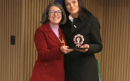 Duas mulheres seguram um troféu nas mãos. Elas estão lado lado, uma vestida com casaco preto e outra de roupa vermelha. Elas sorriem.