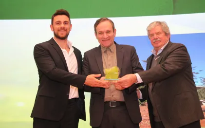 Três homens de terno e camisa social sorriem no palco enquanto o do meio segura um troféu transparente com detalhes verdes.