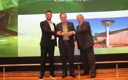 Três homens de terno e camisa social sorriem no palco enquanto o do meio segura um troféu transparente com detalhes verdes.